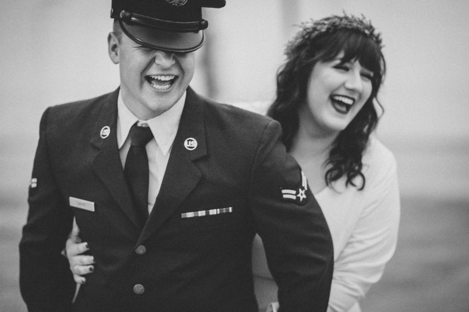 A black and white photograph of a couple laughing together. The man, dressed in a military uniform with a hat tilted slightly forward, grins broadly while the woman, wearing a white dress and a delicate headpiece, playfully holds onto him from behind. Their joyful expressions capture a moment of love and happiness.