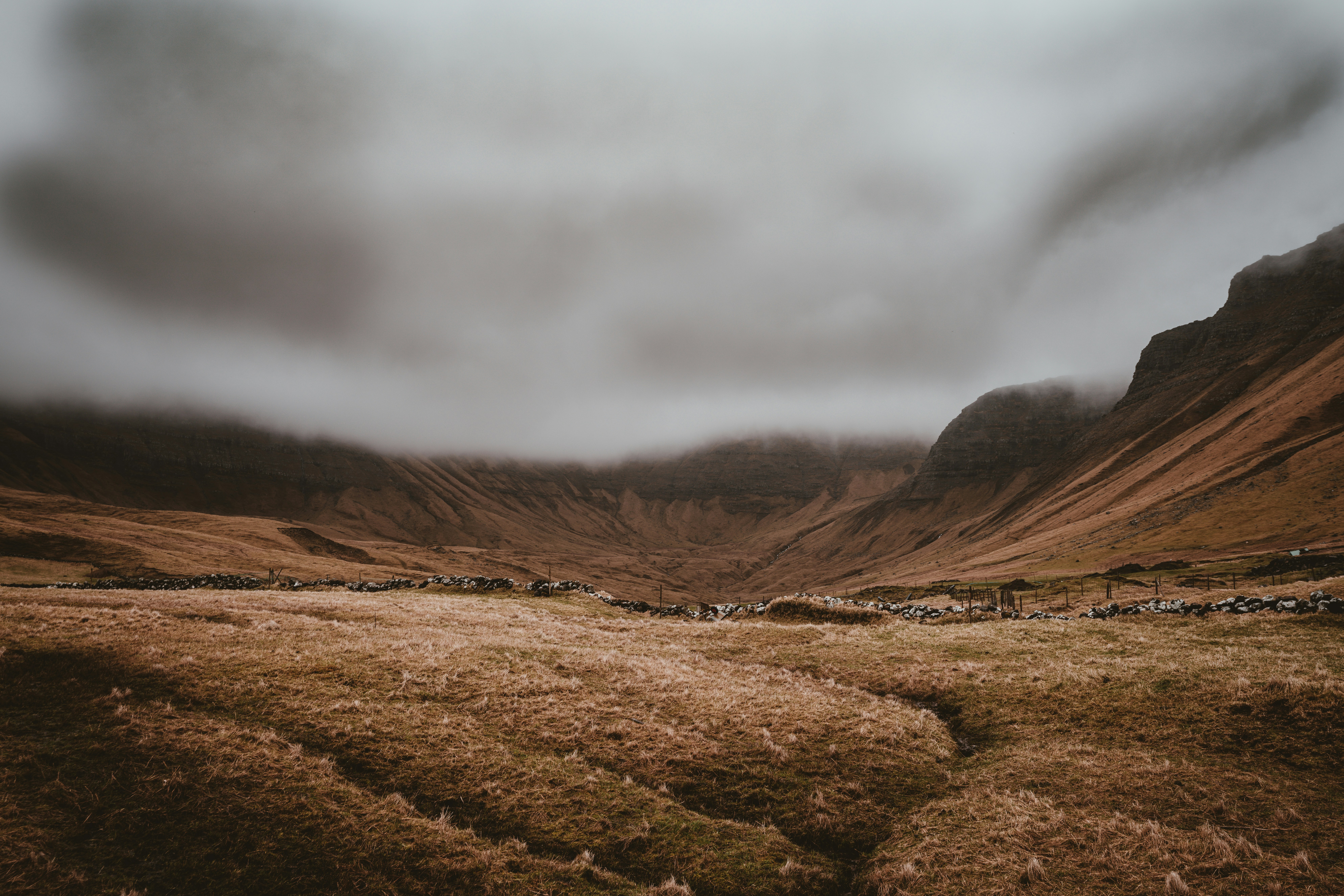 A vast, moody landscape of a rugged valley with steep, barren hillsides under a thick layer of low-hanging clouds. The golden-brown grass in the foreground appears dry and weathered, with a faint path leading toward a stone wall that stretches across the scene. The mountains in the background are partially obscured by mist, creating an atmospheric, almost otherworldly feel. The overall tone of the image is earthy and subdued, with an overcast sky adding to the dramatic and isolated ambiance.