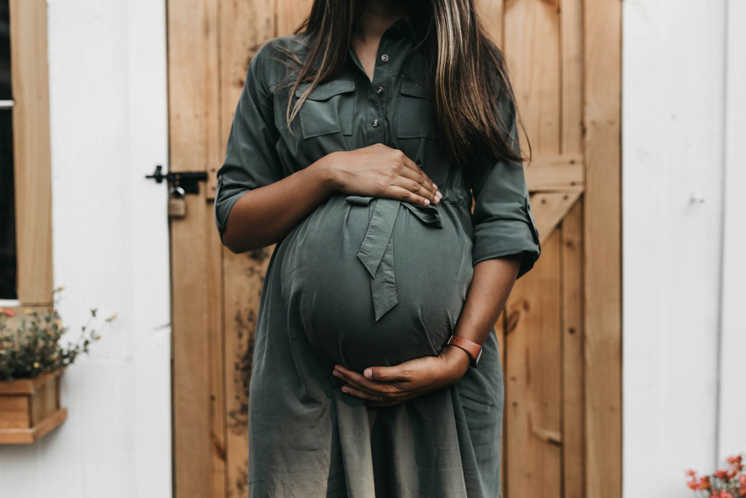 A pregnant woman in a green button-up dress stands in front of a rustic wooden door, gently cradling her baby bump with both hands. The image captures her from the neck down, highlighting her maternity dress, a leather wristband, and the serene atmosphere of the outdoor setting. A small flower box with plants is visible to the left, adding a cozy and natural touch to the scene.