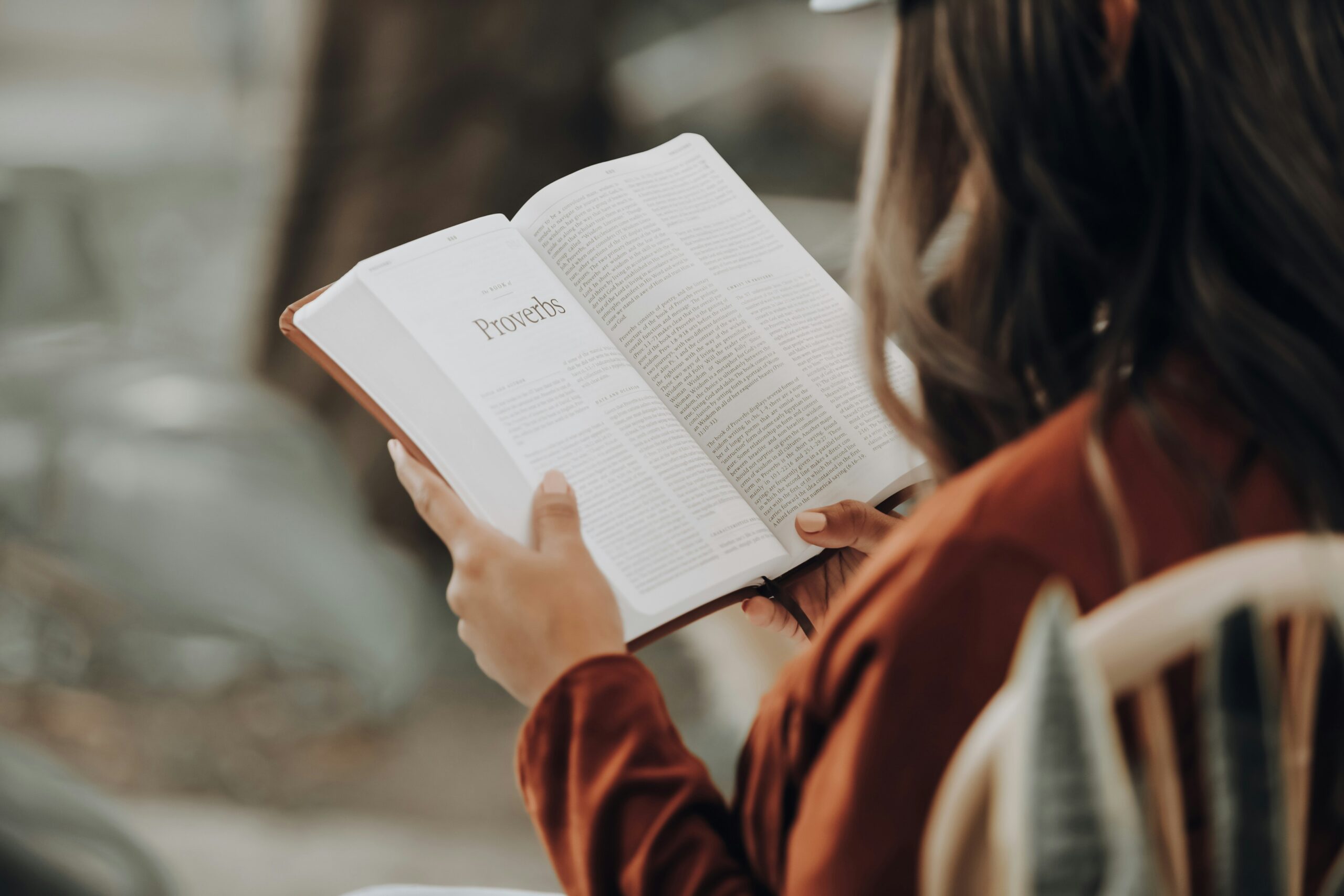 A woman in a brown jacket sits on a wooden chair, reading an open Bible. The page is turned to the Book of Proverbs, and her hands gently hold the book. The background is blurred, creating a peaceful and contemplative atmosphere.