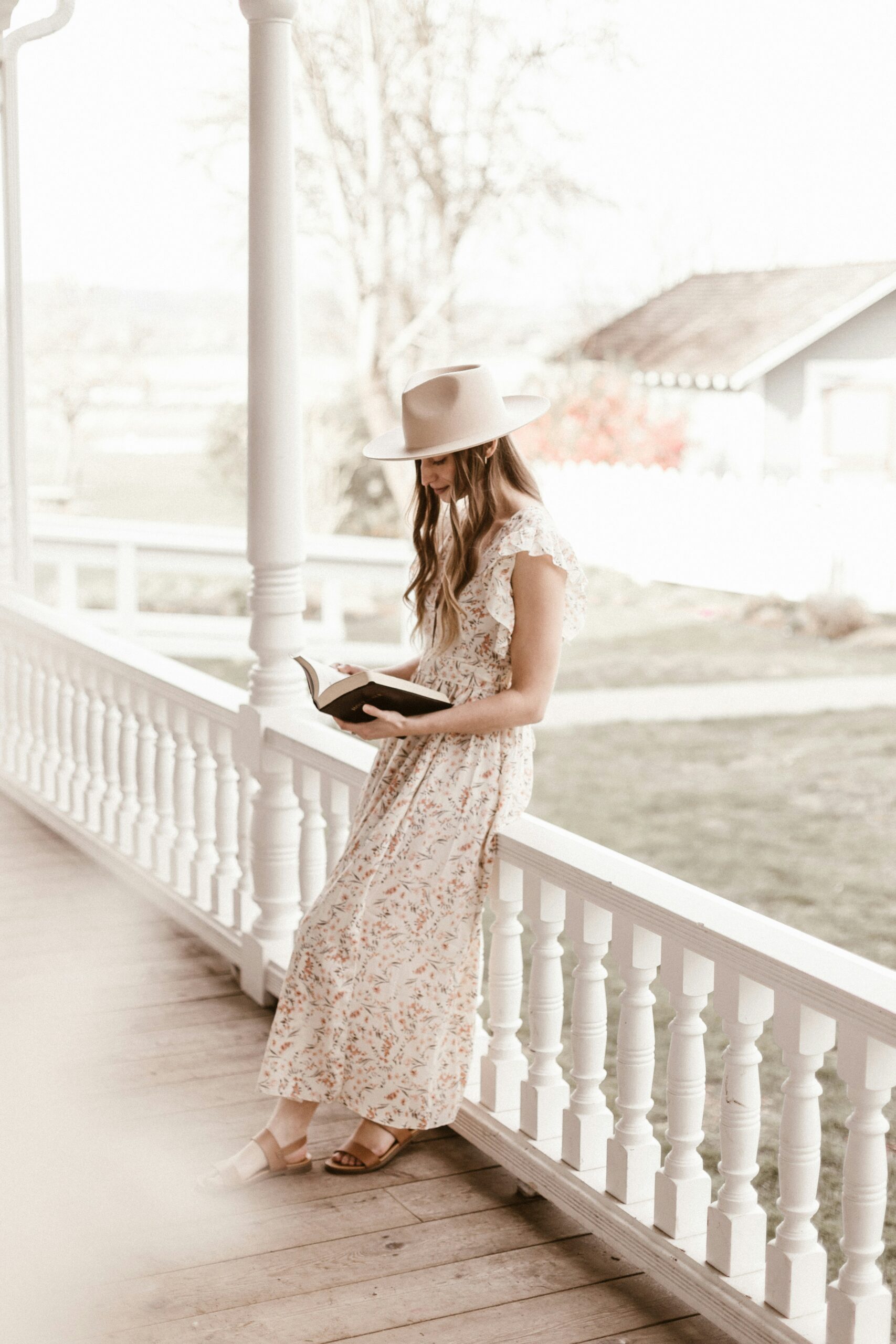 A young woman in a flowing floral dress and a wide-brimmed hat leans against the railing of a white wraparound porch, engrossed in reading a book. The soft, warm lighting and countryside background create a peaceful, timeless atmosphere.